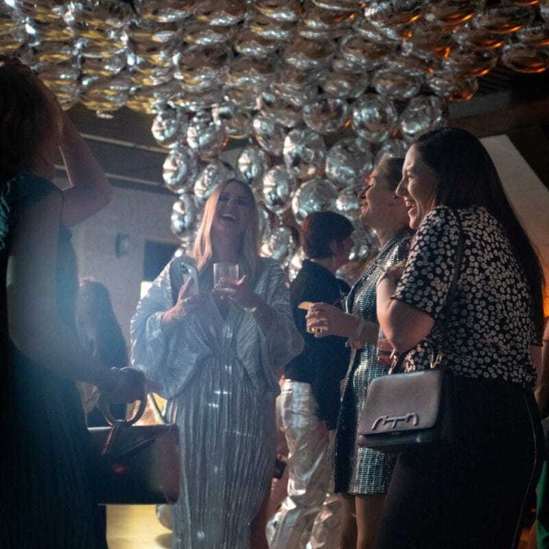 At the MacKenzie Gala, a group of women engage in lively conversation at a party. One woman, dressed in a shiny silver outfit, stands out among the crowd. The ceiling is adorned with numerous reflective silver balloons, enhancing the festive atmosphere.