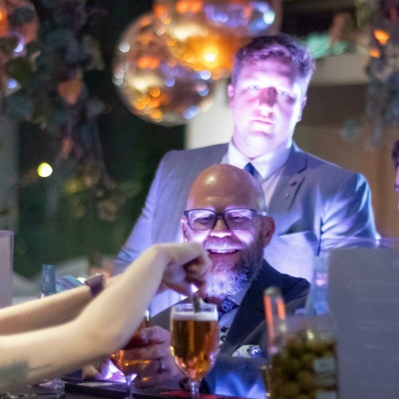 A person with a beard and glasses is smiling at the MacKenzie Gala while a bartender pours a drink at the bar. Another person in a suit stands attentively in the background. The scene is illuminated with soft, colorful lighting and there's a jar of olives on the counter.