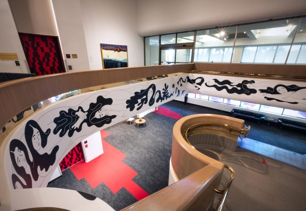 Spiral staircase with a curving banister adorned with bold black and white abstract figures. The staircase leads to an open space with red and gray carpeting, modern art pieces, and a glass-walled meeting room in the background.
