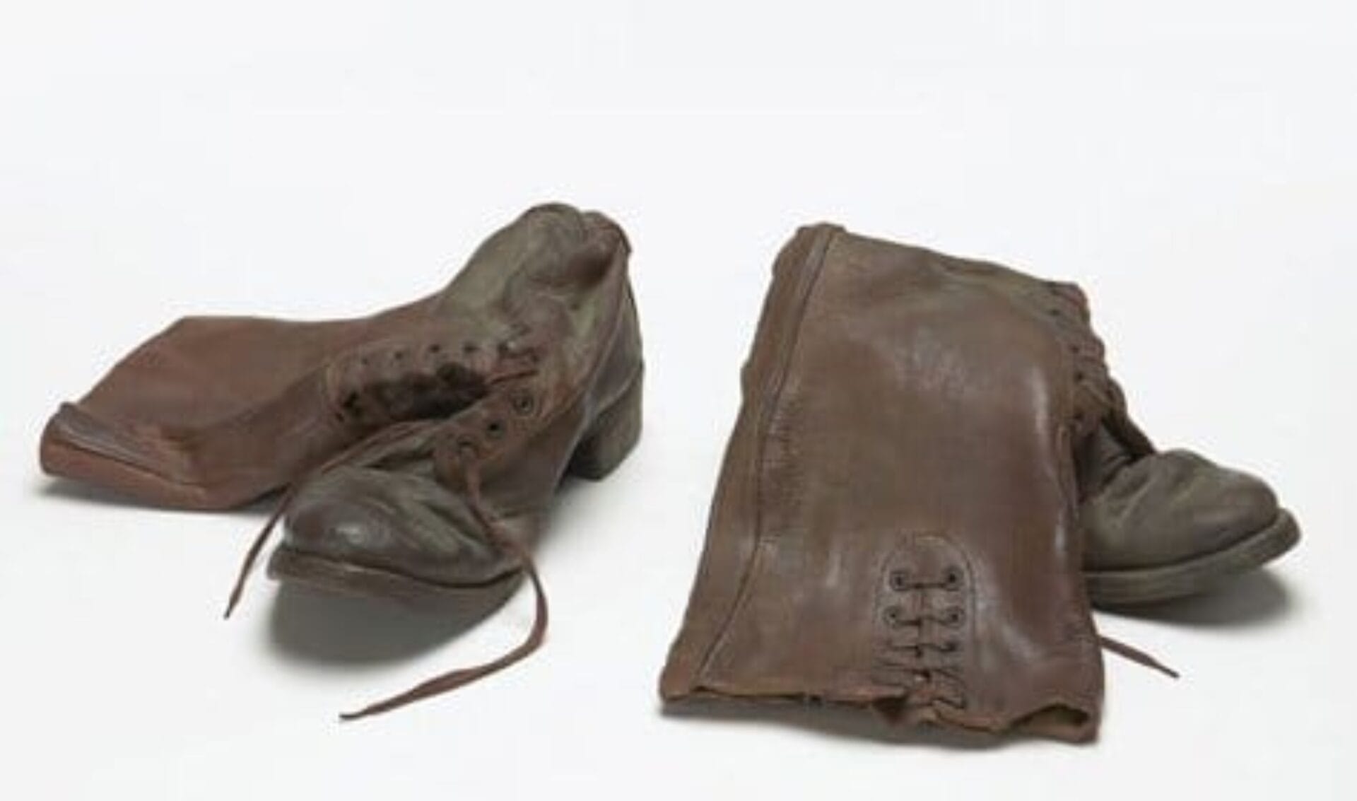 A pair of old, worn, brown leather boots with missing heels and bent shapes placed side by side on a white background. The laces are loosely tied, and the boots show significant signs of wear and age.