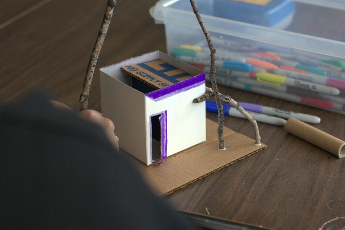 The image shows a small cardboard house with a purple-colored door and window being crafted, reminiscent of a Pinch Pot Portrait. The house is on a piece of cardboard with tree branches surrounding it. Various colored markers and a plastic container with art supplies are visible on the table.