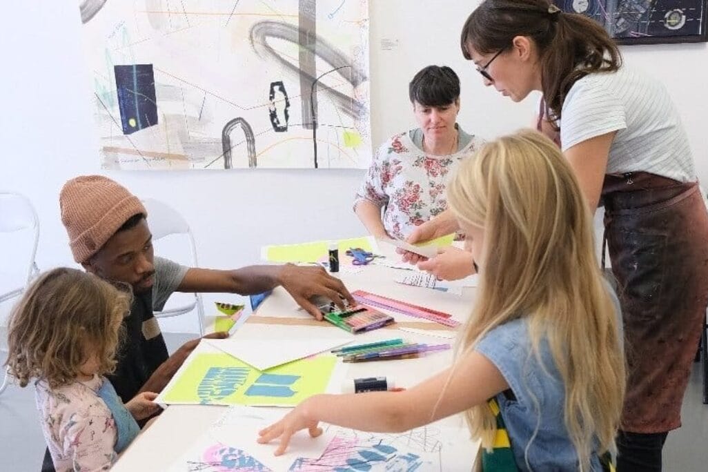 A group of adults and children engage in an art activity around a table. A man and a young girl sit on the left, while two women and another child stand on the right. Various art supplies and colorful papers are scattered on the table, and abstract artwork hangs in the background.