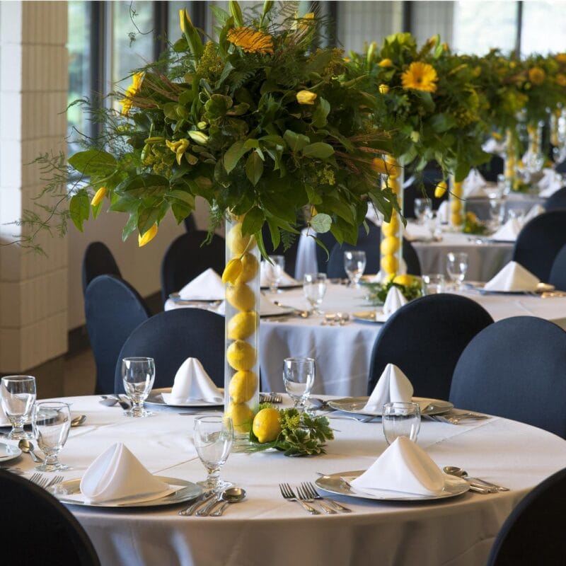 Round tables set out in the Gallery, with table settings laid out, and large centre piece floral arrangements in the middle of each table.