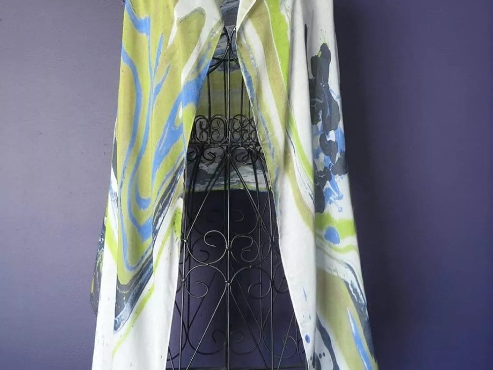 A River Run Scarf by Kay Jay Studio with abstract green, blue, and yellow patterns is draped over a black wrought iron stand against a plain dark purple background. A small tag with a matching design hangs from the scarf.