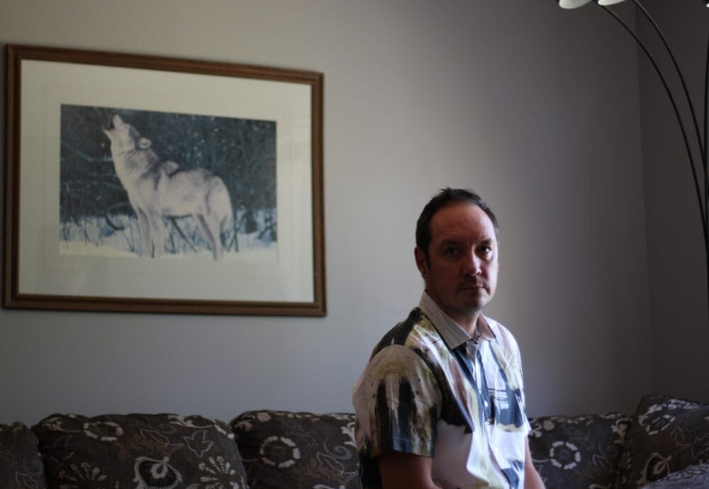A person with short dark hair is seated on a patterned sofa in a dimly lit room. On the beige wall behind them is a framed picture of a howling wolf in the snow. A tall floor lamp is positioned to the right.