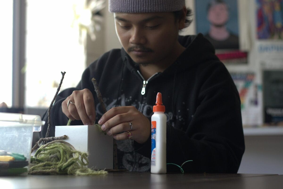 A person wearing a beanie and a hoodie is focused on a craft project. They are holding a twig and applying glue from a bottle to it. Various crafting materials, including yarn, more twigs, and even some supplies for their upcoming Pinch Pot Portrait, are scattered on the table in front of them.