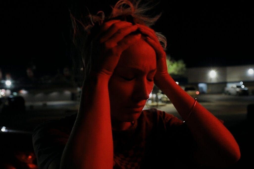 A person stands in a dimly lit area at night, holding their head in distress with both hands. Their expression is pained, and the scene is illuminated by red light, creating a dramatic and intense atmosphere reminiscent of the spinning chaos of a pinwheel.