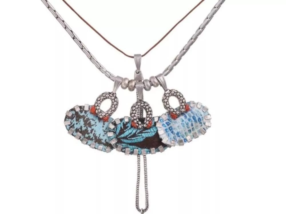 A Necklace by Ayala Bar featuring three oval-shaped pendants with blue and black patterns, adorned with beaded details. The center pendant has a small hanging tassel with a blue beaded accent. The chains include a silver-tone link and brown cord.