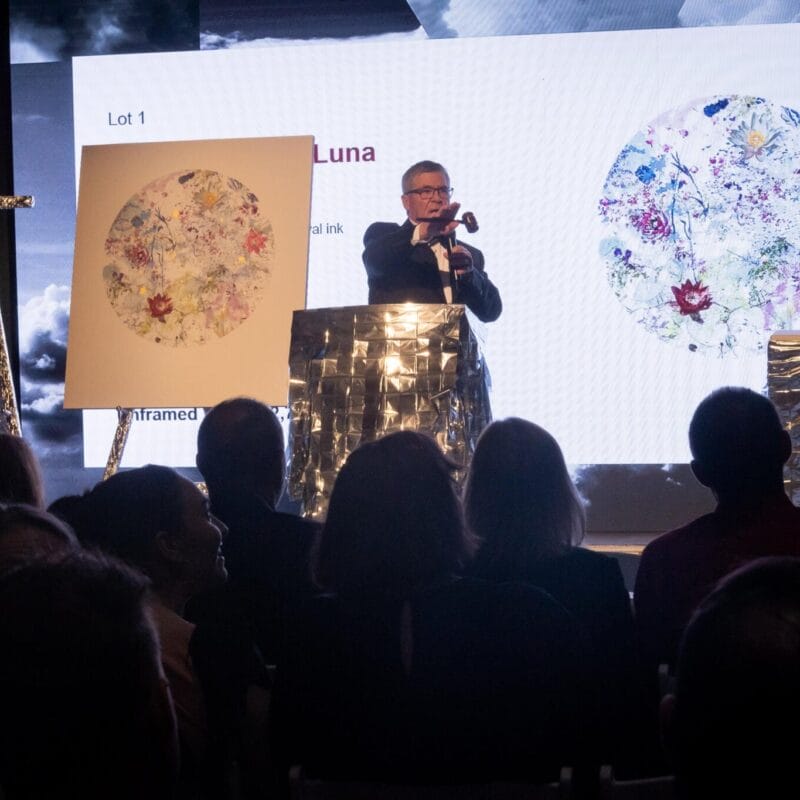 An auctioneer stands behind a podium with a gavel, presenting a circular artwork displayed on an easel next to him. The details of the art piece are projected on a large screen behind him. At the prestigious MacKenzie Gala, the audience is seated attentively, focused on both the auctioneer and the artwork.