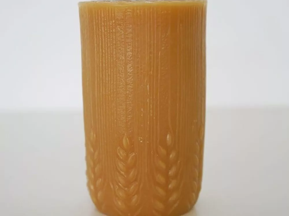 A Wheat Pillar by Joan's Beeswax Candles with a textured surface, featuring vertical lines and a wheat motif at the base, standing on a plain white background.