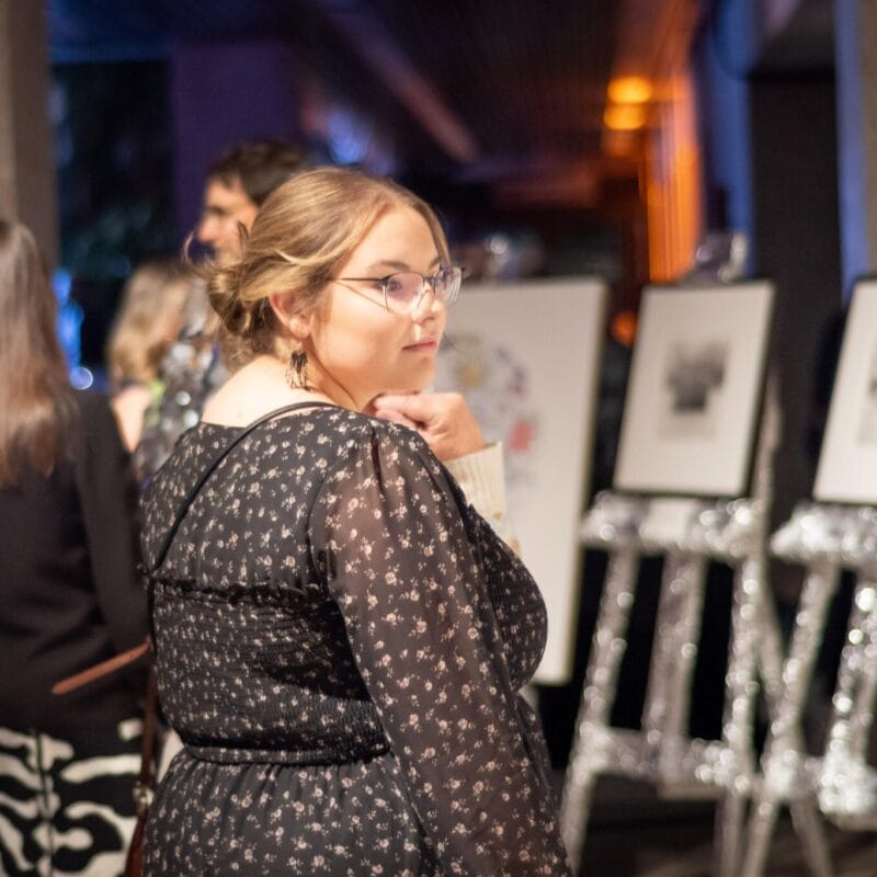 A woman with glasses and a dark floral dress looks over her shoulder at an art exhibit during the MacKenzie Gala. Framed artworks on easels are visible in the background, with several other people viewing them. The setting appears to be a gallery or event space with dim lighting.