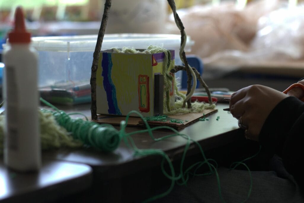A person is working on a small, colorful model house with yarn and glue on a table. Among the various craft supplies—green string, a glue bottle, and a clear plastic container—is also a Pinch Pot Portrait showcasing their artistic flair. The person's hands are visible as they meticulously work on the project.