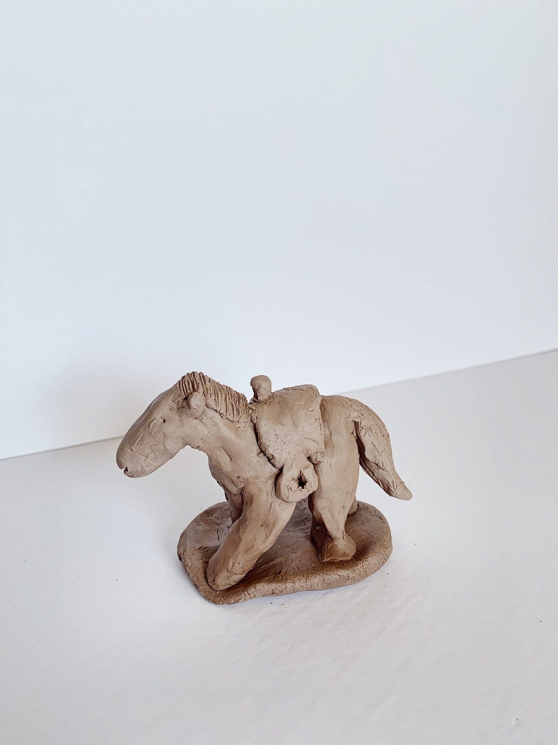 A small, hand-sculpted clay figurine of a horse standing on a flat base. The horse has a rider on its back with basic features. The sculpture is unpainted and has a simple, rustic appearance. The background is plain white.