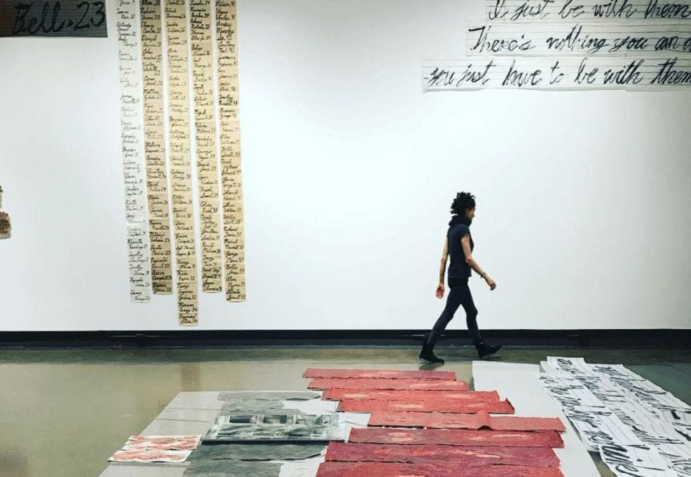 A person walks through an art installation featuring various canvases laid on the floor, including red textured pieces and black-and-white prints. In the background, large sheets with handwritten text are displayed on the wall. The room has a minimalist, industrial feel.