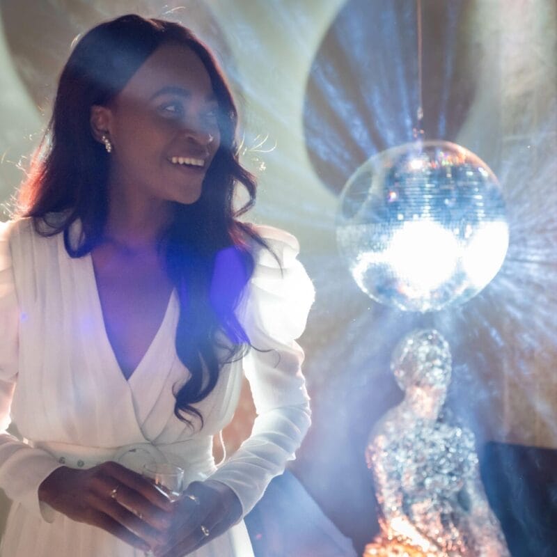 MacKenzie Gala, in a white dress, stands smiling under a disco ball. Light reflections create a vibrant atmosphere in the room. She holds a small glass in one hand while her long hair cascades over her shoulders.