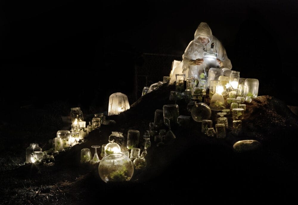 A person tending to small plants being grown in make shift mini green houses. The greenhouses each have a small lamp casting light in the darkness.