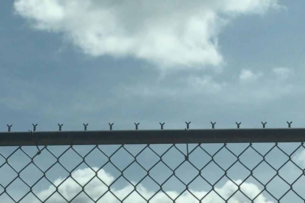 A chain-link fence topped with small loops is silhouetted against a blue sky with scattered white clouds. The fence extends horizontally across the image, creating a contrast between the metal structure and the sky.