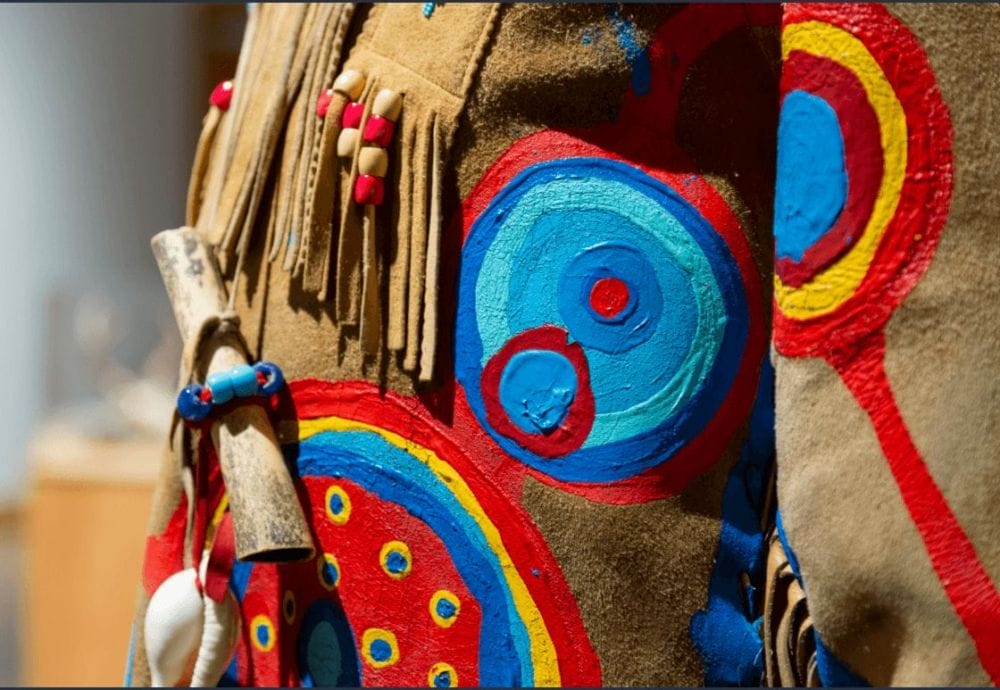 Gros plan d'un tissu en daim beige orné de motifs peints circulaires et vibrants en rouge, bleu et jaune. Le tissu présente des franges en perles et un élément en bois noué avec des perles bleues. Les décorations lui donnent un aspect artisanal et complexe.