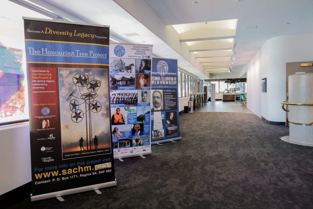 Two standing banners are displayed in a brightly-lit hallway. The banners promote community projects and services with text and images, including a tree sculpture and various people. The hallway has white walls and large windows, allowing natural light to fill the space.