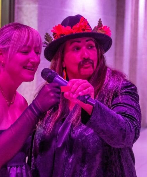Two people are singing together using a shared microphone. The person on the left is wearing a strapless dress, long gloves, and a red hair accessory. The person on the right has a long mustache, beard, and is wearing a hat with red leaves, both lit by pink lighting.