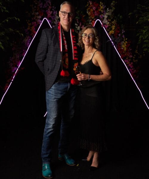 A man and a woman pose in front of a dark backdrop with purple neon triangle lights. The man wears a checkered blazer, jeans, a scarf, and glasses, while the woman is in a sleeveless black dress and glasses, holding a drink. Colorful flowers can be seen in the background.