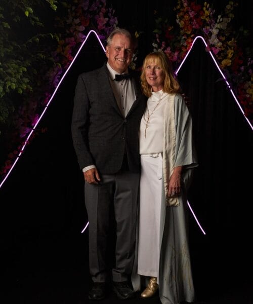 A man and woman pose together in formal attire against a black backdrop adorned with neon lights and floral arrangements. The man wears a dark suit and bow tie, while the woman wears a light-colored outfit and overcoat. They both smile at the camera.