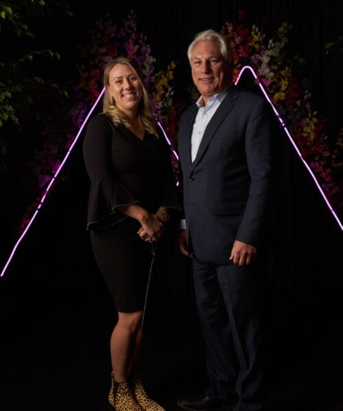 A woman in a black dress and leopard print shoes stands next to a man in a dark suit, both smiling. They are in front of a black backdrop with a colorful triangular light display and floral decorations behind them.