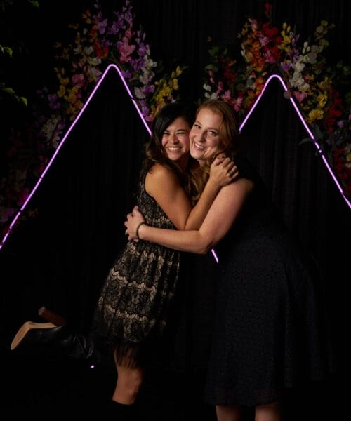 Two women pose together, smiling and hugging, in front of a black backdrop adorned with neon pink and white lights forming triangle shapes. The background also features colorful floral decorations and greenery on either side. Both women are wearing dresses.