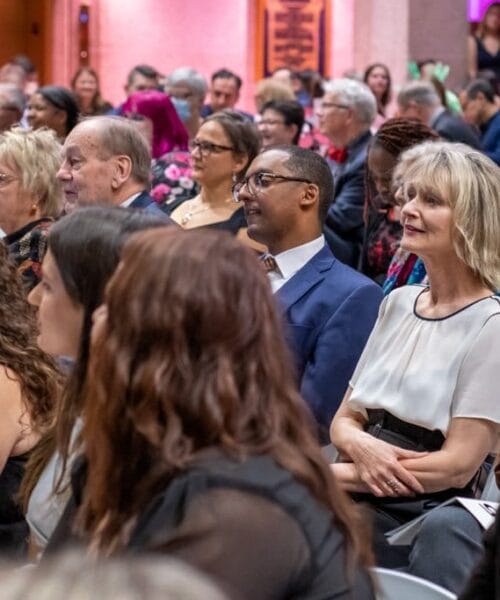 A diverse group of people sit closely together in an indoor setting, attentively listening to a speaker. The audience includes individuals of various ages, genders, and ethnicities. The background has warm lighting and some potted plants. Everyone appears engaged.