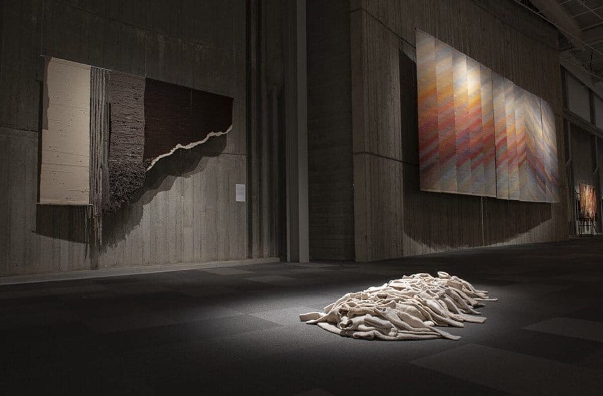 Small sweaters are piled one on the other in the middle of the gallery floor. Two large textile pieces are hung on the wall in the background.
