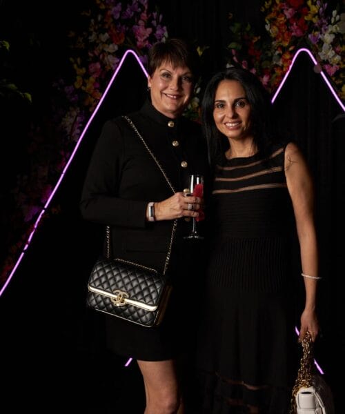Two women pose together in front of a backdrop adorned with flowers and neon pink triangular lights. They are dressed in elegant black outfits, with one holding a drink and the other carrying a gold handbag. The scene is framed with greenery on the sides.