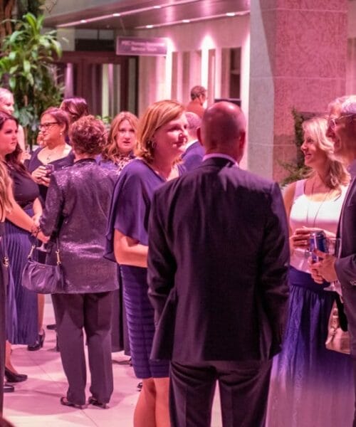 A group of people in formal attire engaging in conversation and drinks at a well-lit indoor event with pink lighting. Some are gathered in small groups, mingling and chatting. A few people are standing near a bar or refreshment area. Decor includes plants and artwork.