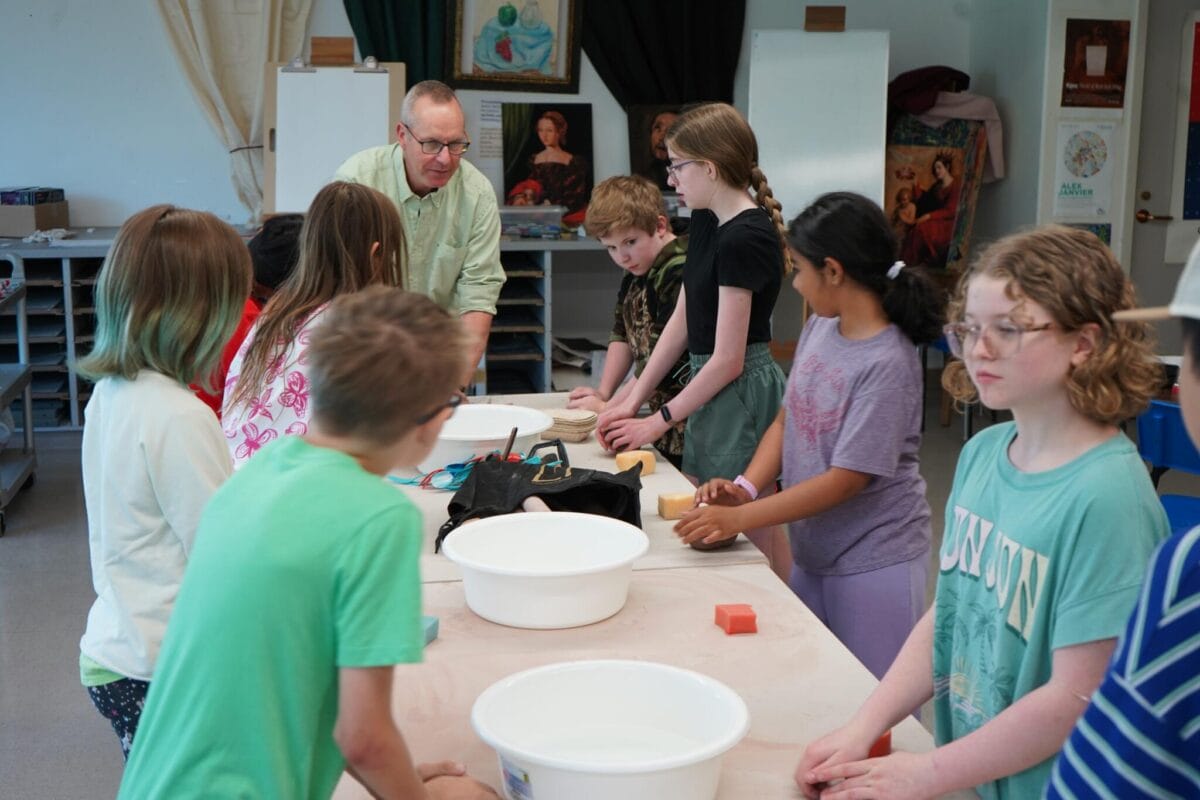 Un enseignant se tient à une table entouré d'élèves, démontrant une activité avec des bols d'argile et d'eau dans une salle de classe remplie de fournitures d'art et de peintures.