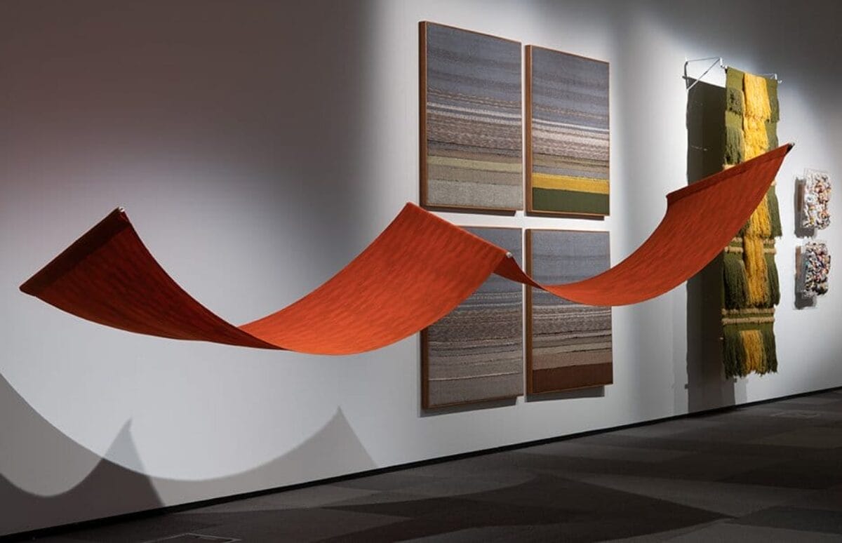Red textile artwork hanging on display as if it's flying through the air horizontally. Other textile works can be seen in the background hanging on the wall.