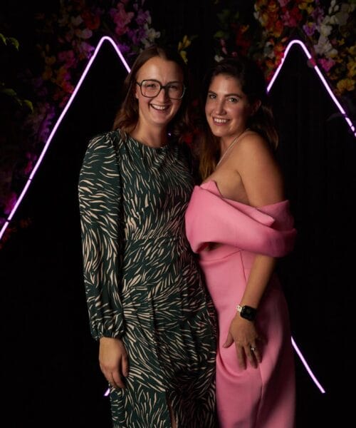 Two women stand close together, smiling, against a dark backdrop with pink neon triangle outlines. One wears a green patterned dress and glasses, the other a pink off-shoulder jumpsuit. There are plants and floral decorations around them.