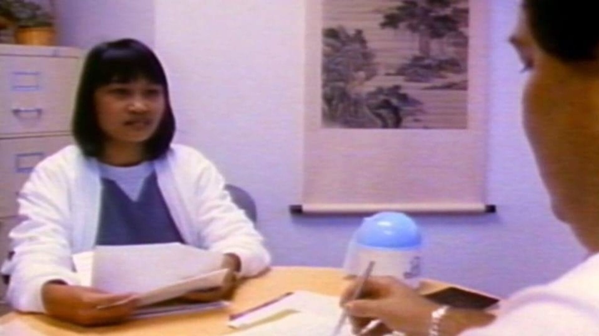 A woman in a white coat holds papers while talking to a seated person in the foreground, who is writing. They are in an office with a filing cabinet and a wall hanging in the background. A blue and white container and a clipboard are on the table.