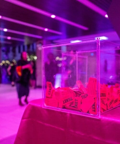 A transparent box filled with red raffle tickets sits on a pink-covered table in a room lit with purple lighting. A glass of drink and a laptop are also on the table. In the background, people are mingling near a round, black-clothed table.