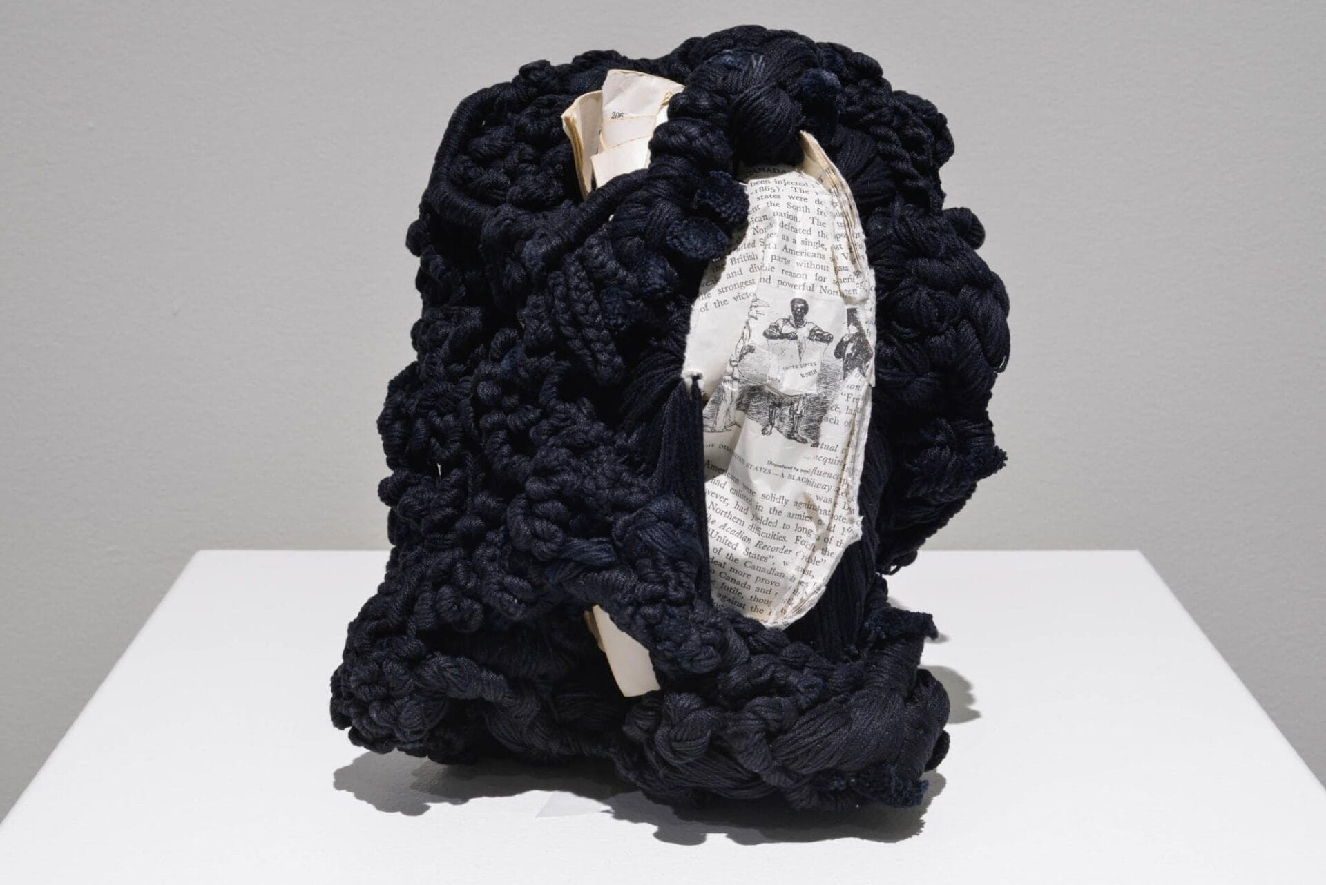 A sculpture featuring a roughly textured, dark woven material enveloping pieces of white paper with printed text and images. The abstract form stands on a flat white surface against a light grey background.