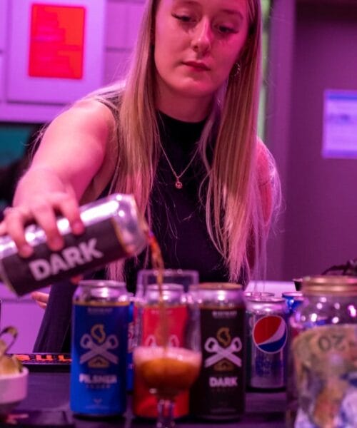A person with long blonde hair pours a beer labeled 