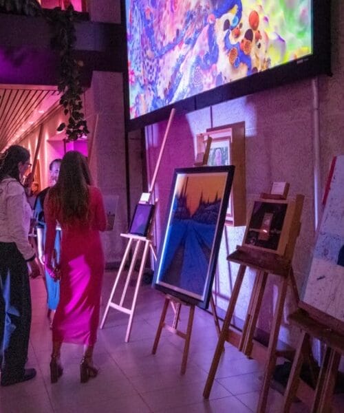 A group of people standing in an art gallery, observing paintings on easels. Some paintings are abstract, while others depict landscapes. The gallery is illuminated by pink and purple lights. The atmosphere is lively and the attendees appear engaged in conversation.
