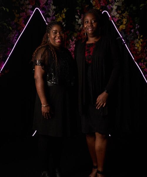 Two women stand side by side, smiling, in front of a dark backdrop adorned with colorful flowers and purple neon triangle shapes. They are both wearing black outfits and have long braided hairstyles.
