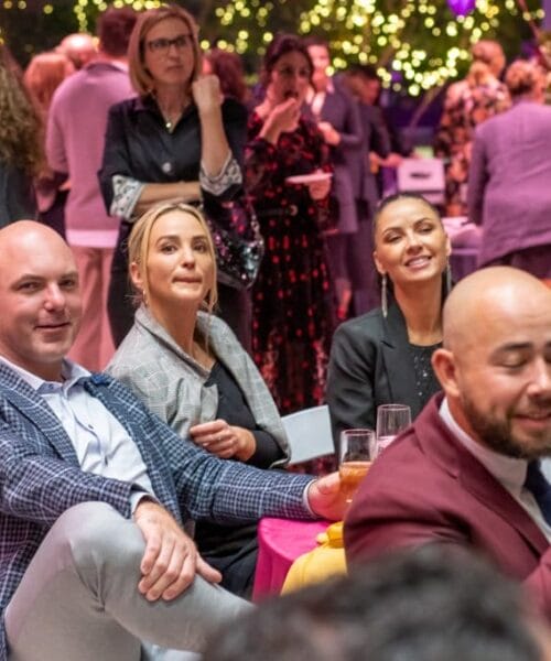 A group of people seated together in an indoor event with festive lighting and decorations. Some individuals are smiling and holding drinks, while others appear engaged and attentive. The background shows more attendees mingling and socializing.