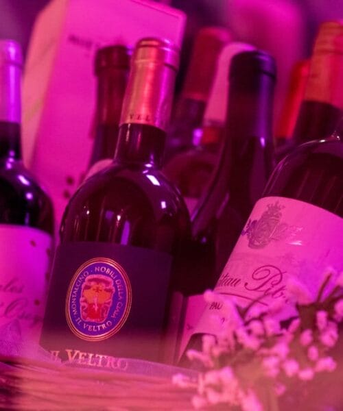 A wicker basket filled with various bottles of wine, each adorned with different labels. The image is enveloped in a pinkish-purple hue, adding a dramatic flair to the scene. Small decorative flowers are tucked around the bottles, enhancing their presentation.