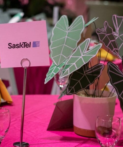 A dining table is decorated with pink tablecloth, yellow napkins, wine glasses, and a centerpiece featuring paper leaves in pastel colors. A sign on the table reads 