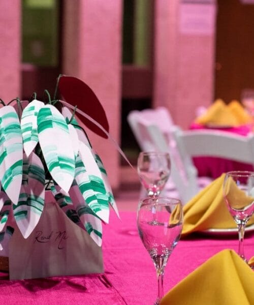 A dining table is adorned with a red tablecloth and yellow napkins folded into triangular shapes. At the center is a white and green paper leaf decoration. The background shows more similarly set tables and a standing person in a brightly lit room.