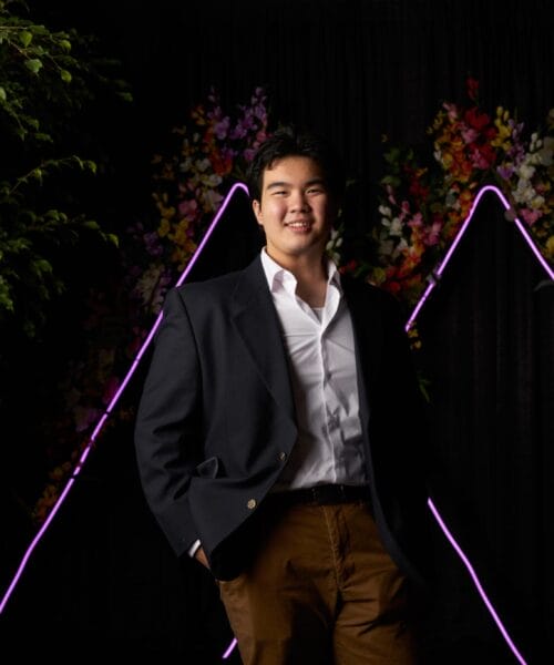 A man in a dark blazer and brown pants stands in front of a dark background decorated with triangular neon lights and colorful flowers. He is smiling and has one hand in his pocket. Green foliage frames the scene on both sides.