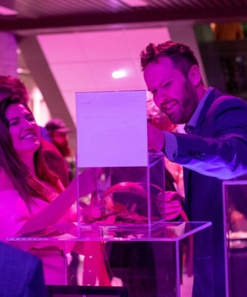 A group of people is gathered around a display case at an event. A man in a suit and a woman in pink clothing are engaged in conversation, looking at the exhibit. The lighting is predominantly purple, creating a vibrant atmosphere. Other attendees are in the background.