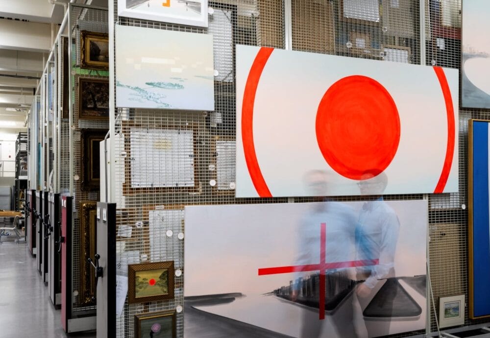 A storage area filled with various artworks, including large canvases with abstract and geometric designs. Two figures, blurred due to motion, walk through the space like whispers from the vault. The artworks are displayed on metal racks, and some smaller framed pieces are also visible.