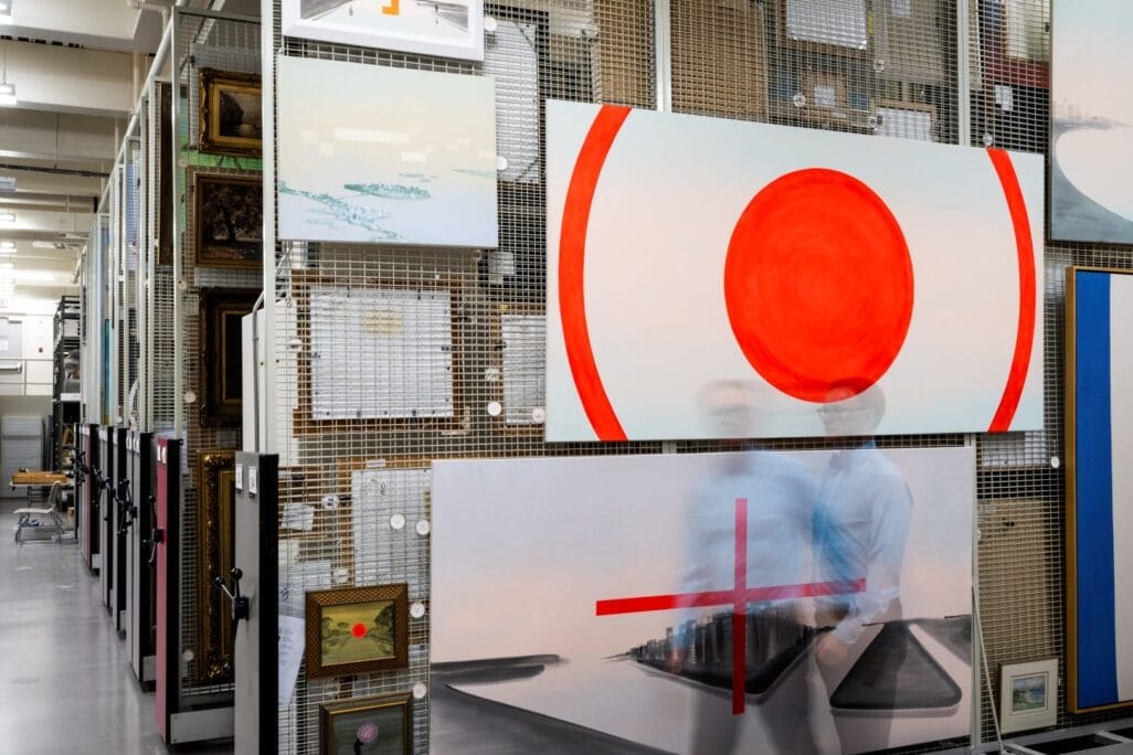 A storage area filled with various artworks, including large canvases with abstract and geometric designs. Two figures, blurred due to motion, walk through the space like whispers from the vault. The artworks are displayed on metal racks, and some smaller framed pieces are also visible.