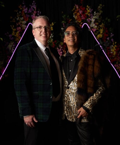 Two people posing together in front of a floral backdrop with purple neon lights. The person on the left wears glasses, a green plaid jacket, and a tie, while the person on the right has a leopard print jacket, a fur stole, and glasses.